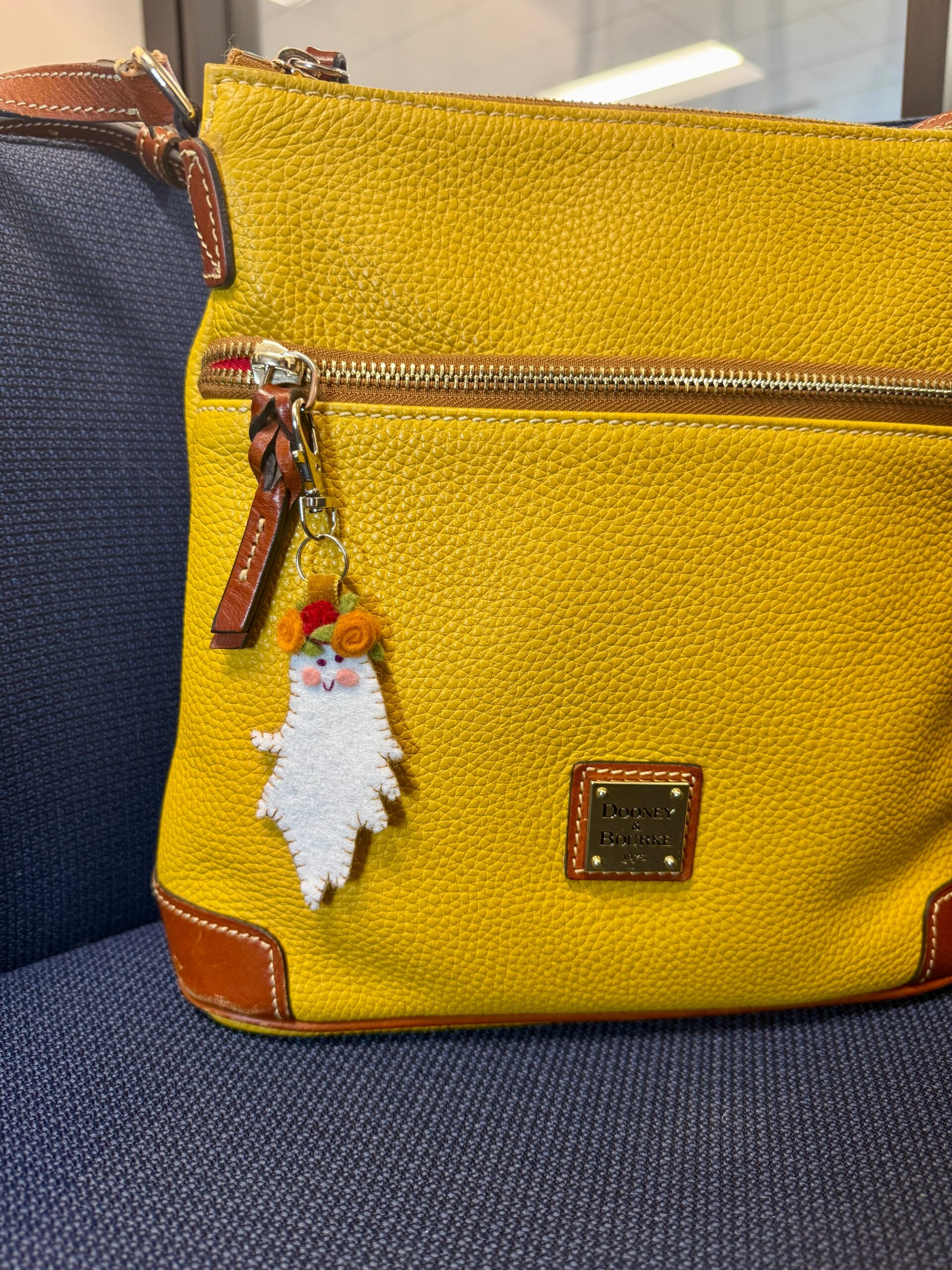 Boo-quet felt ghost charm with an orange and red flower crown clipped to the zipper of a yellow leather bag