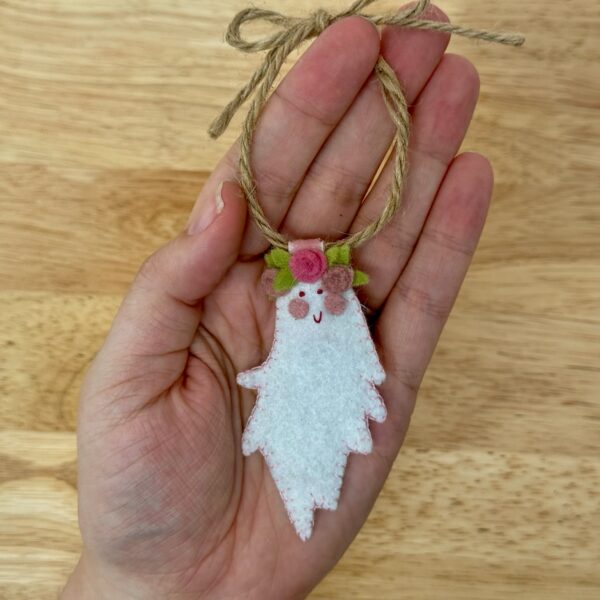Finished Boo-quet felt ghost ornament with a pink spiral rose flower crown, hanging from a twine loop held in a hand