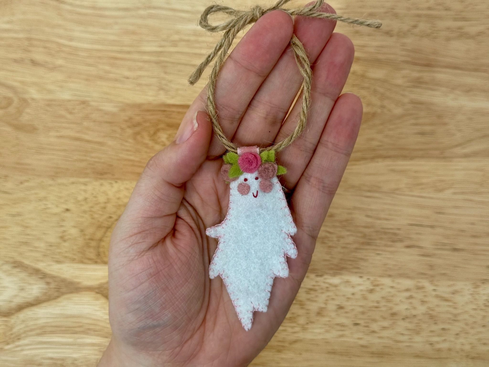 Finished Boo-quet felt ghost ornament with a pink spiral rose flower crown, hanging from a twine loop held in a hand