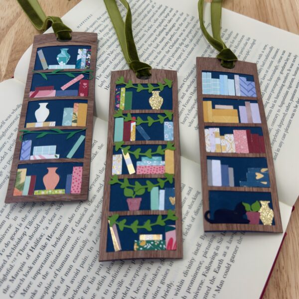 Three finished mini bookshelf bookmarks with wooden frames, colorful paper books, and green velvet ribbons resting on an open book