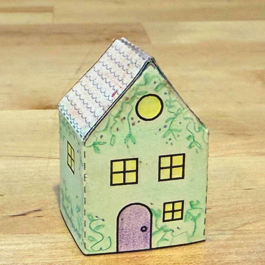 A small paper house colored with pencils, green with a purple door, standing on a wooden counter
