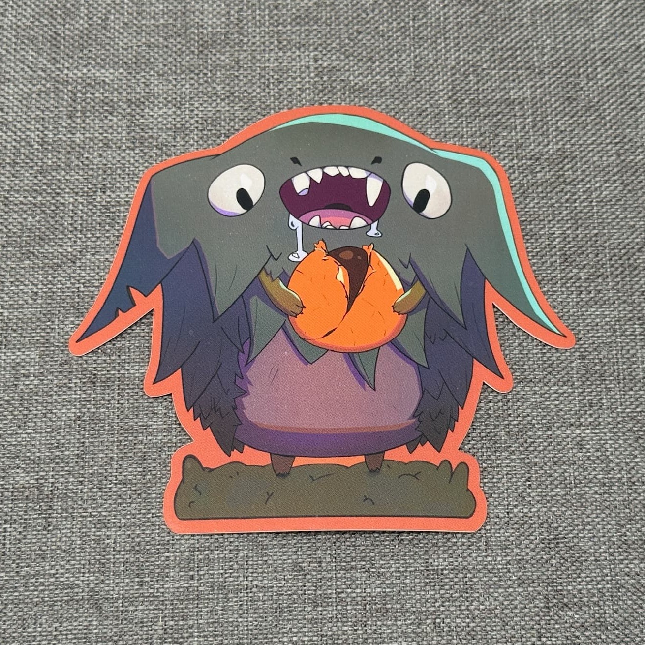 Devon the Candy Thief sticker: a dark feathered creature with wide eyes and fangs, clutching a piece of orange candy, outlined in orange