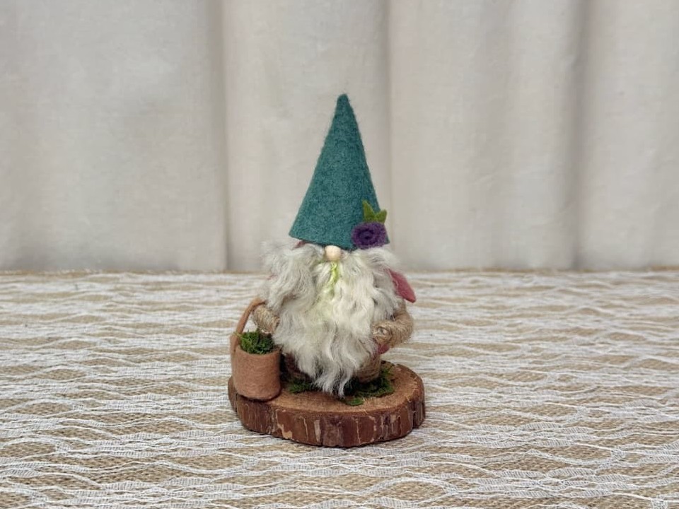 Earl, a handmade gnome with a teal felt hat, purple felt rose, moss in his white yarn beard, and a tan felt basket of moss on a wood slice base