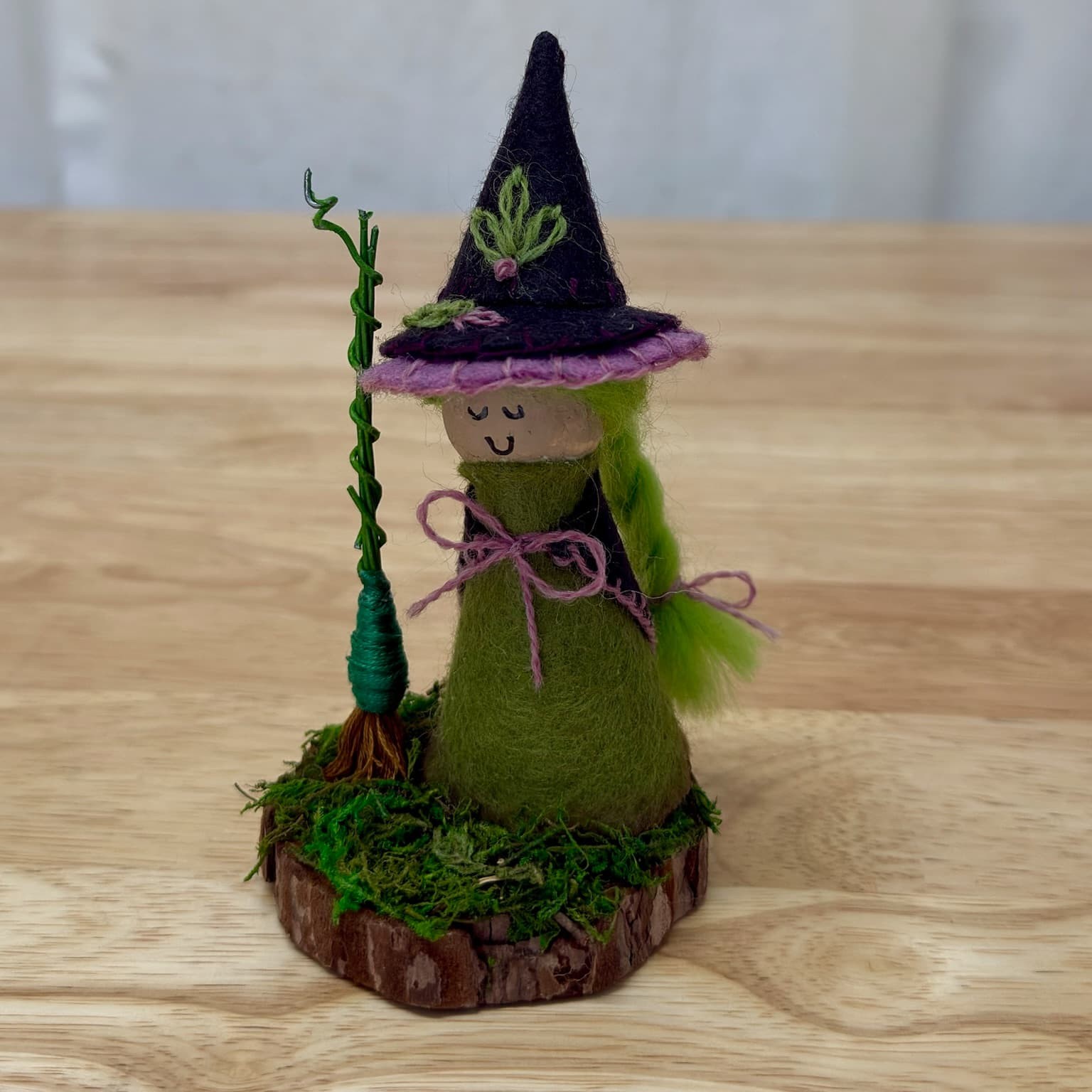 Handmade little witch figurine with a pointed felt hat and a broom, standing on a wood-slice base