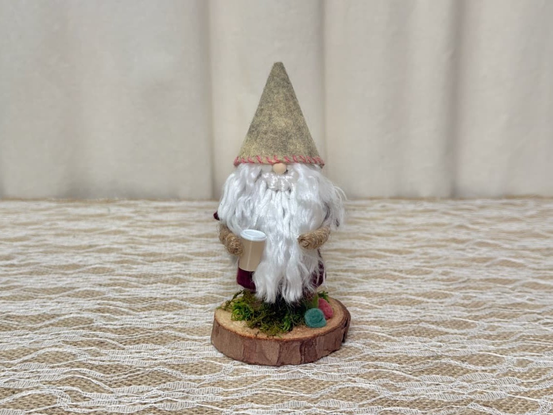 Zak, a handmade gnome with an oatmeal felt hat, white yarn beard, burgundy coat, holding a tiny coffee cup on a wood slice base